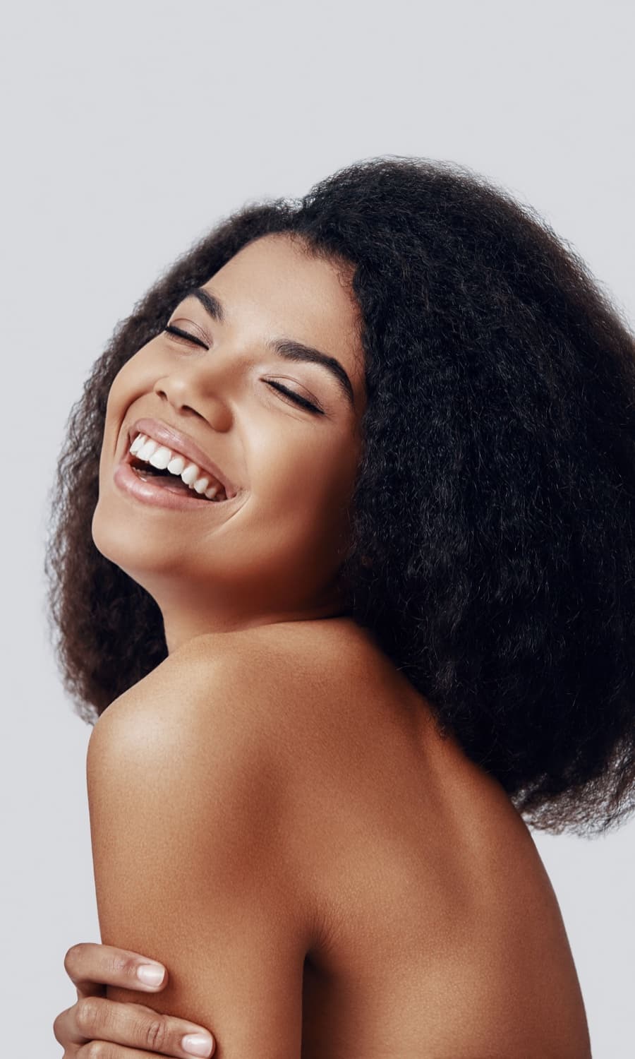 Woman with curly hair laughing, eyes closed, against a light backdrop
